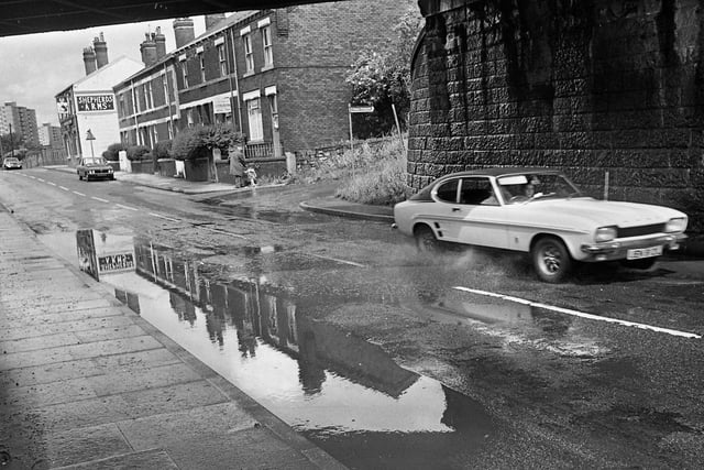 A classic Ford Capri splashes through a flooded section of road under the railway bridge on Warrington Road, Lower Ince, in the summer of 1975. In the background is the Shepherds Arms pub.